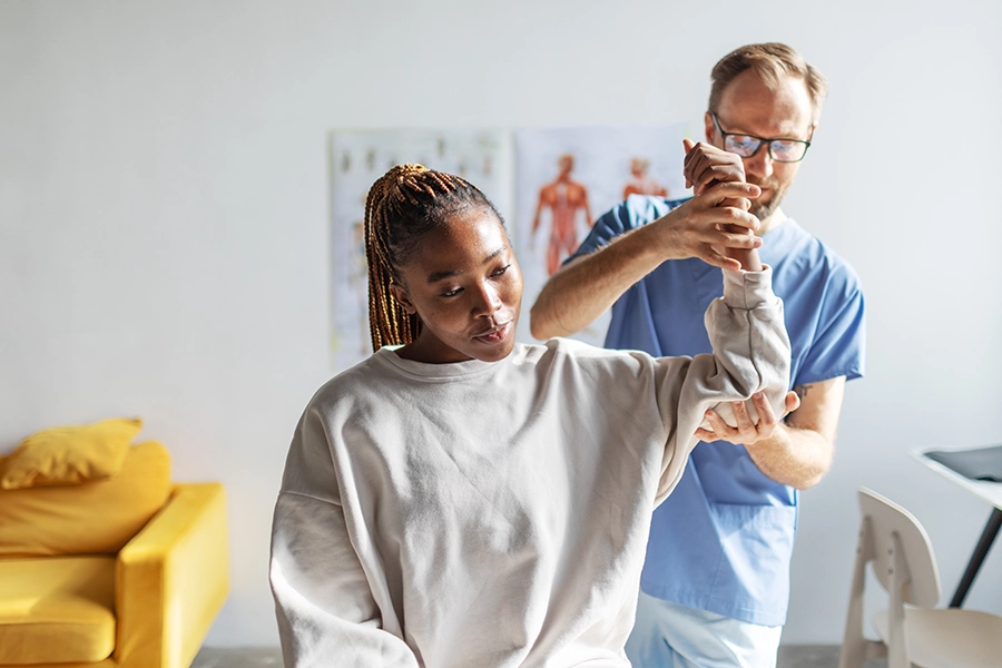 Physical therapist in blue scrubs supporting a seated patient's arm during a shoulder range-of-motion exercise in a clinic room.